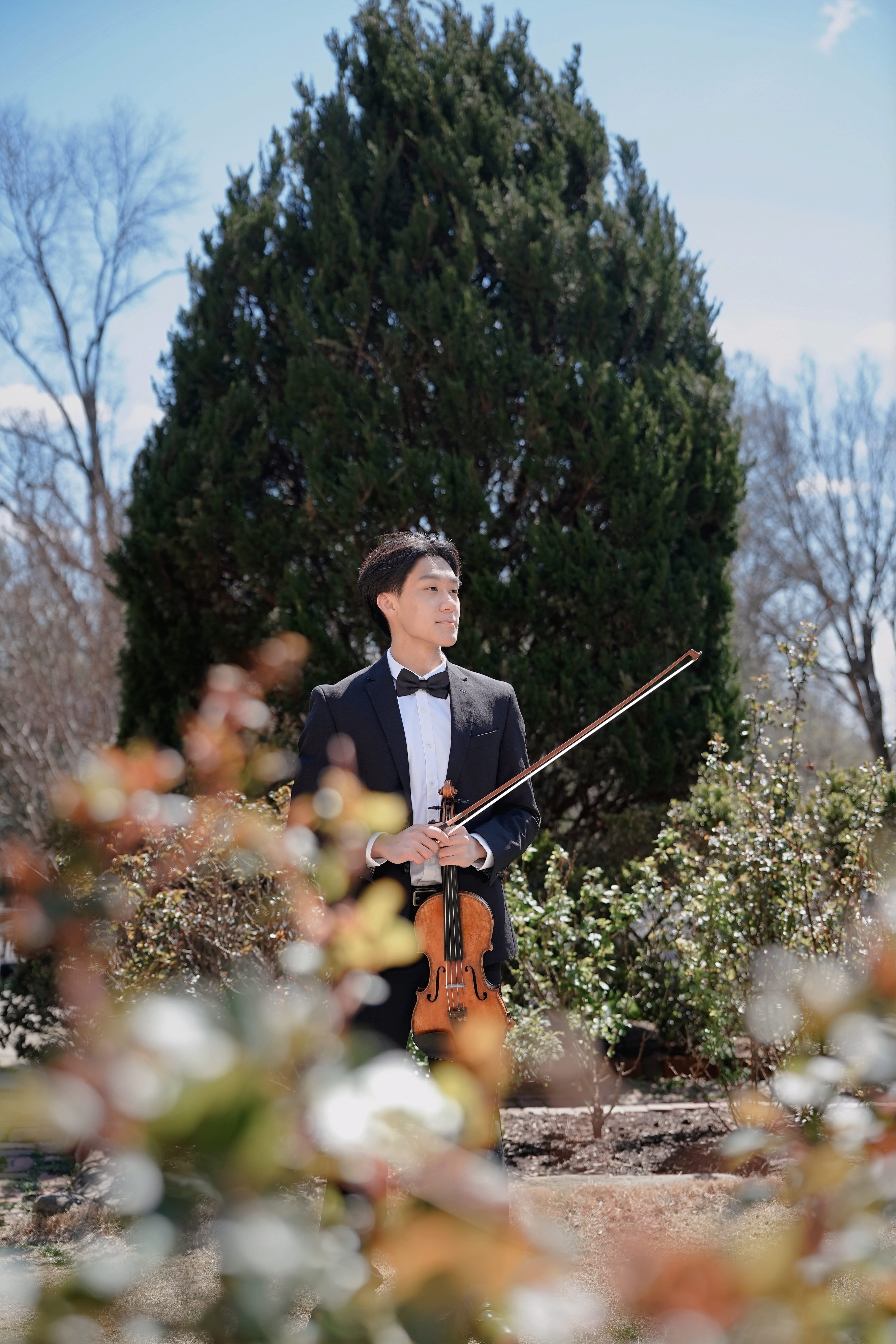 Bernie Sheng in a black tuxedo holding his violin in a garden